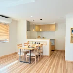 木目の造作ダイニングテーブルが置かれた明るいダイニング。食器が並ぶテーブルと、木製キッチンが調和したナチュラルな空間