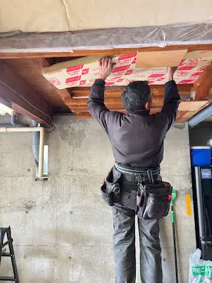 作業員がガレージの天井に断熱材をはめ込んでいる様子。むき出しの梁の間に断熱材を押し込み、周囲には工具や配管が見える施工中の空間