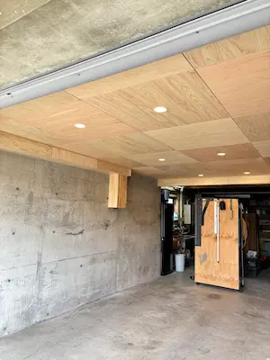 木の天井にダウンライトが整然と並び、明るく仕上がったガレージ内部の様子。コンクリート壁と木の質感が調和し、手前にはキャスター付きの収納ラックが置かれている
